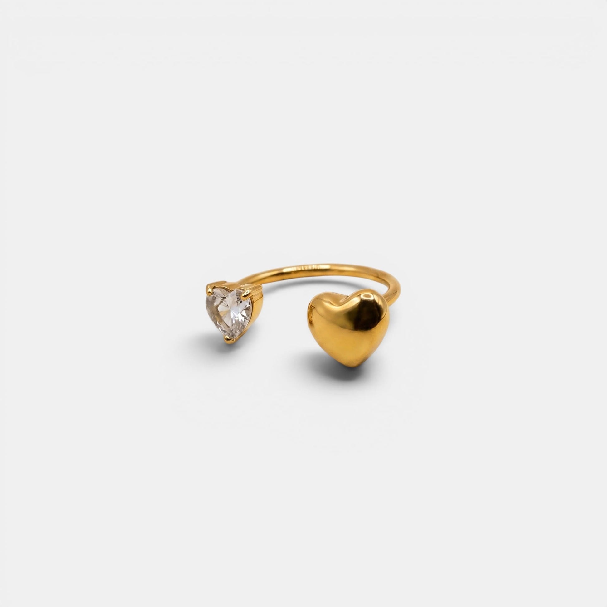 Goldener Amara Heart Ring mit glattem Herz und funkelndem Kristallherz auf cremefarbenem Hintergrund.