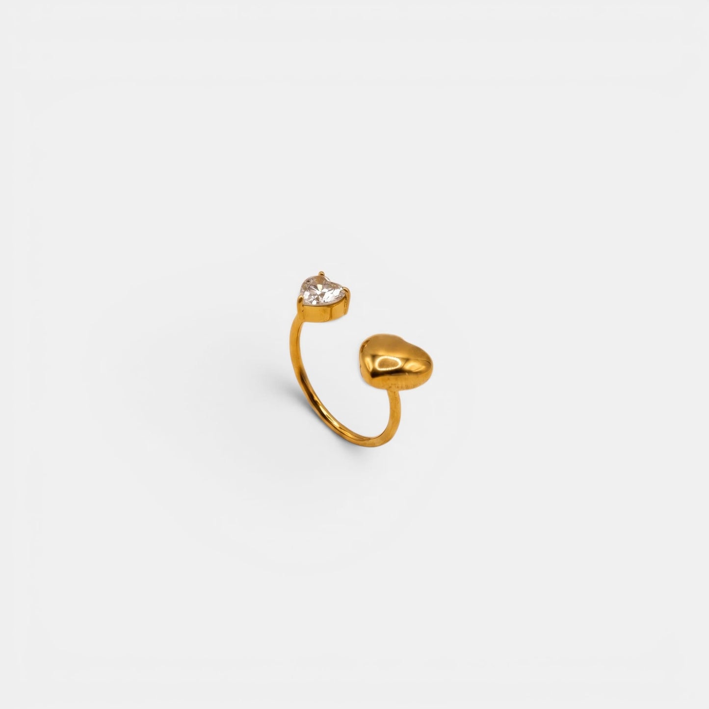 Goldener Amara Heart Ring mit glattem Herz und funkelndem Kristallherz auf cremefarbenem Hintergrund.